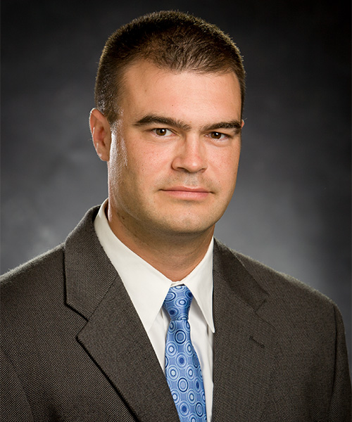 Portrait of a man wearing a dark suit, white shirt and blue patterned tie, facing the camera against a dark studio background.