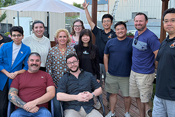 Group photo of 11 people smiling and posing together on an outdoor deck, with two people seated in front and nine standing behind them. A building, canopy and greenery are visible in the background.