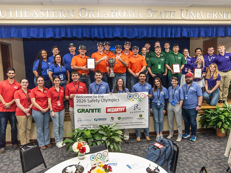 A large group of students and faculty pose on stage holding trophies, certificates, and a banner welcoming attendees to the 2026 Safety Olympics, highlighting team participation and award winners at the event.