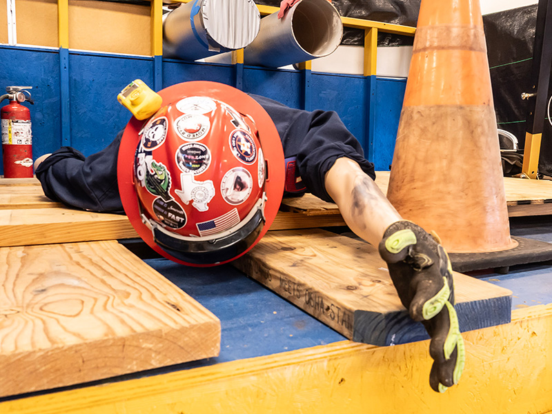 A participant wearing a safety helmet and gloves crawls beneath wooden obstacles during a simulated rescue or confined‑space challenge, demonstrating hands‑on safety training in a competitive exercise.