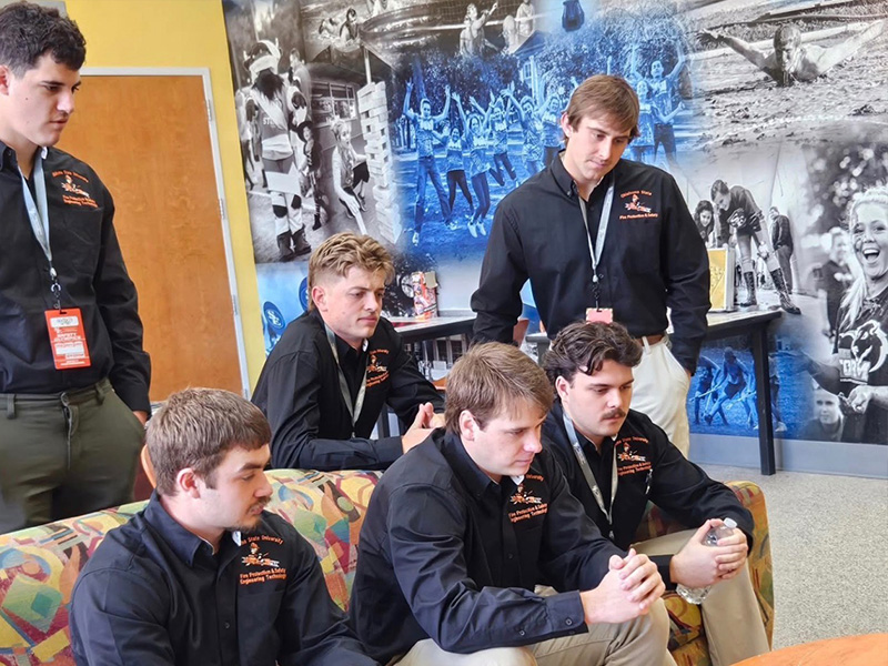 A group of students wearing matching shirts sit closely together watching and discussing a scenario, illustrating team collaboration and problem‑solving during a Safety Olympics preparation or competition activity.