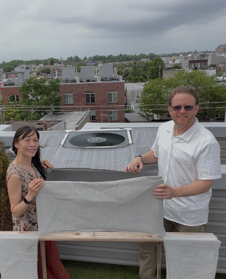 Two people stand on a rooftop next to an air conditioning unit, holding and displaying a fabric cover designed to shade the unit.