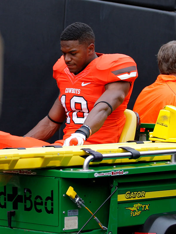 Football player in an orange Oklahoma State Cowboys uniform seated on a medical cart at the sideline during a game.