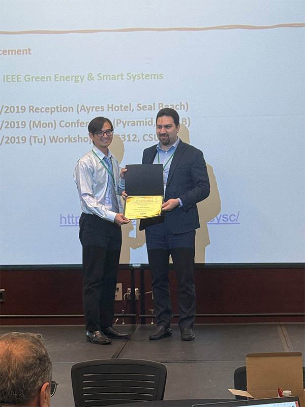 Dr. Hamid Pouya and one of his students stand on a stage to receive a certificate during a conference of the Institute of Electrical and Electronics Engineers.