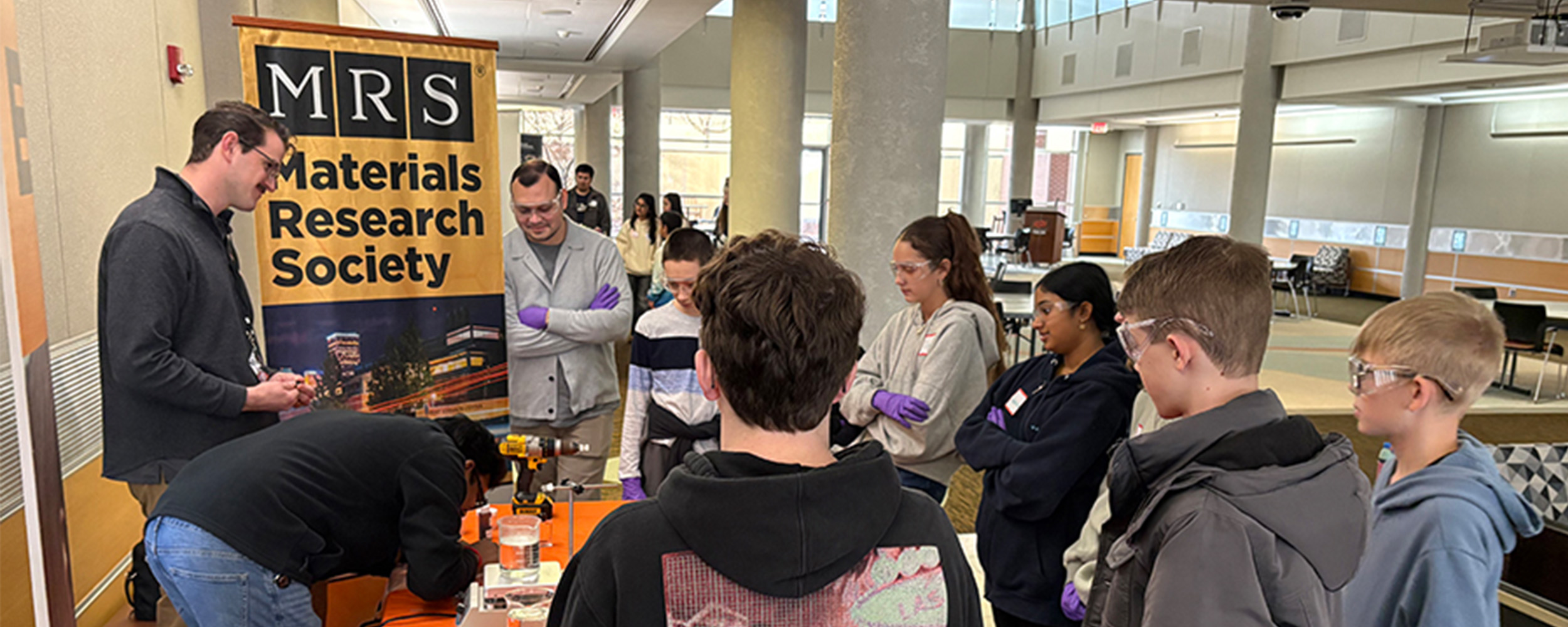 A group of students and instructors gather around a table during a hands‑on materials science demonstration inside a campus building, with a Materials Research Society banner displayed behind them.