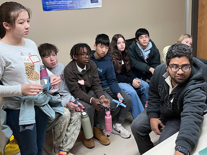 A group of students sit closely together in a classroom while an instructor gestures toward a surface out of frame, indicating participation in a hands‑on educational or research activity.