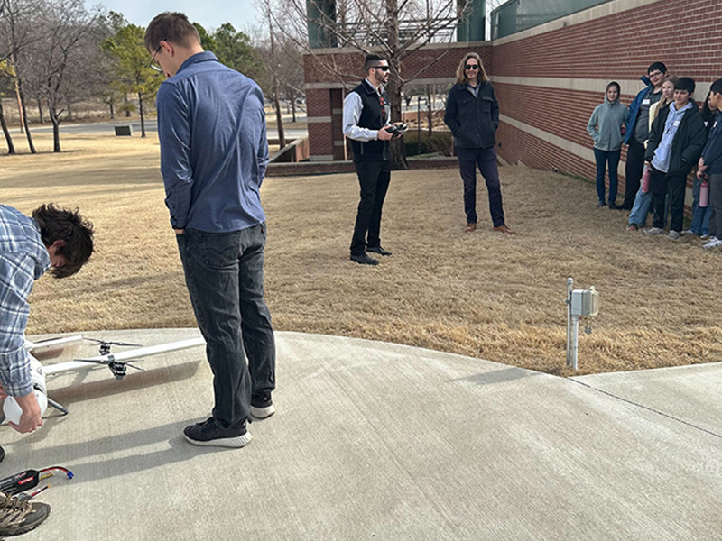 A group of students stand outdoors on a campus lawn watching an instructor demonstrate equipment on a paved area near a building, indicating a hands‑on educational activity.