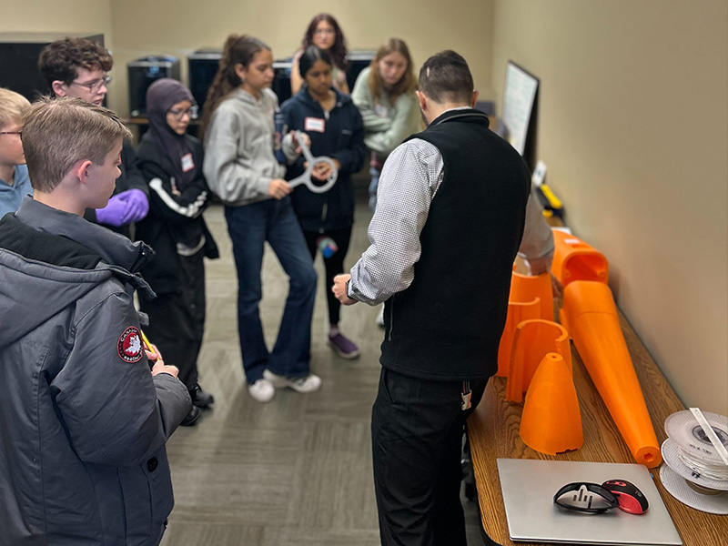 Students stand around a table indoors while an instructor demonstrates materials using orange plastic components, illustrating a hands‑on materials science learning activity.