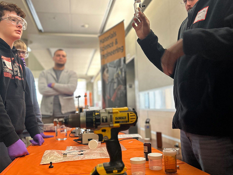 An instructor holds up a small sample vial while students wearing safety goggles observe a hands‑on materials science demonstration at a table with tools and sample containers indoors.