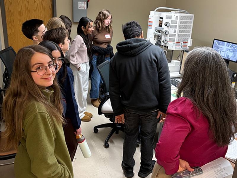 A group of students gather around laboratory equipment and computer monitors indoors while an instructor demonstrates scientific imaging or materials analysis, indicating a hands‑on learning activity.