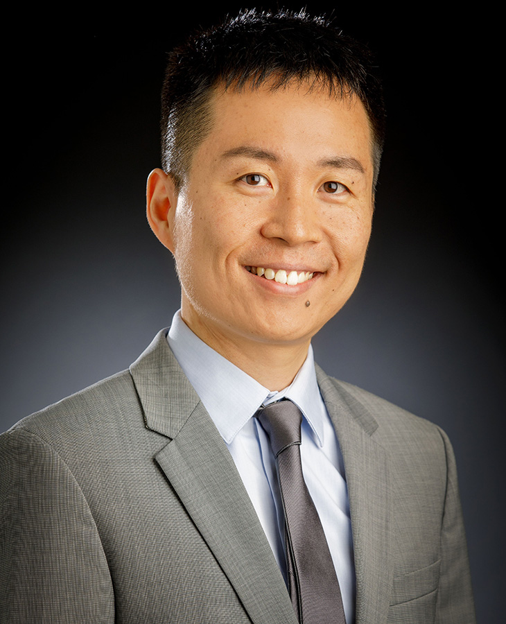 A man with black hair, wearing a brown suit jacket poses for a professional headshot