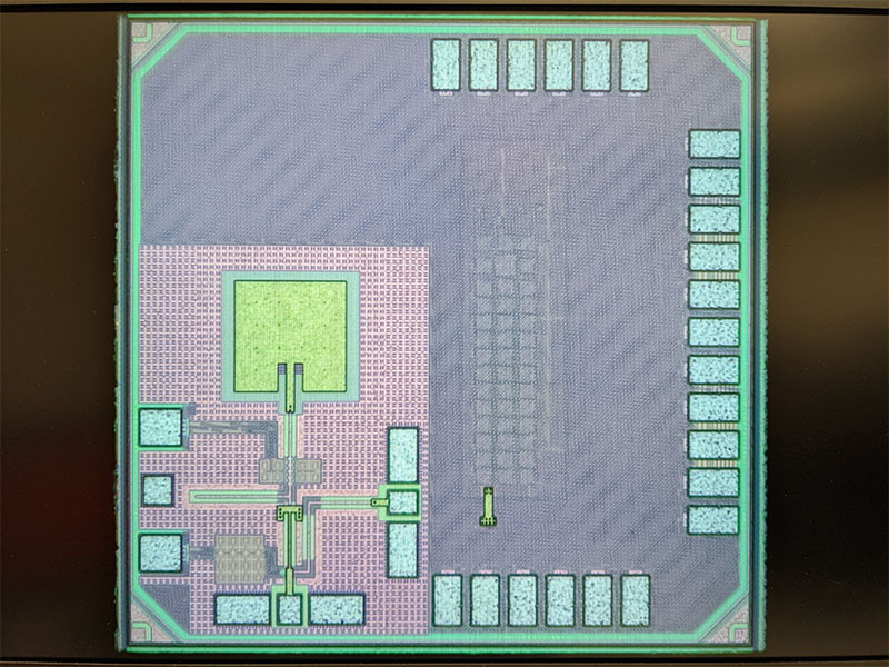 Close‑up micrograph of an integrated circuit showing a square chip with visible circuitry patterns and contact pads around the edges, illustrating the internal layout of a semiconductor device.