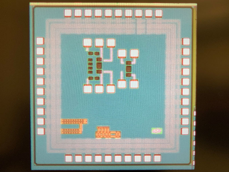 Close‑up micrograph of an integrated circuit showing a square chip with visible circuitry patterns and contact pads around the edges, illustrating the internal layout of a semiconductor device.