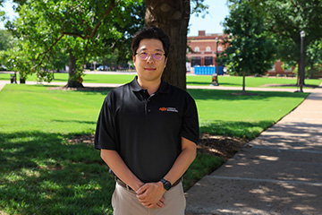 A person wearing a black Oklahoma State University Chemical Engineering polo shirt stands with hands clasped on a shaded campus walkway