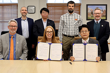 A group of professionally dressed people sit and stand near a conference room table, two people display signed contracts.