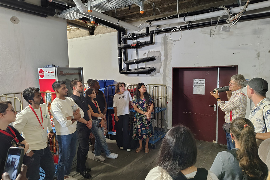 A group of approximately 15 people gathers in an unfinished utility or service room.