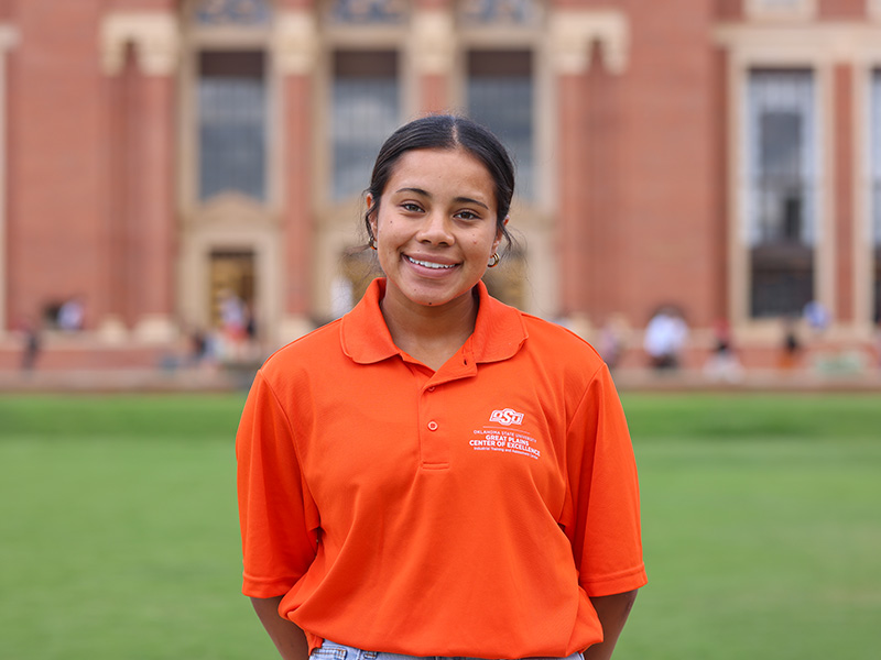 A young woman in a bright orange polo poses on the lawn of Edmon Low library.
