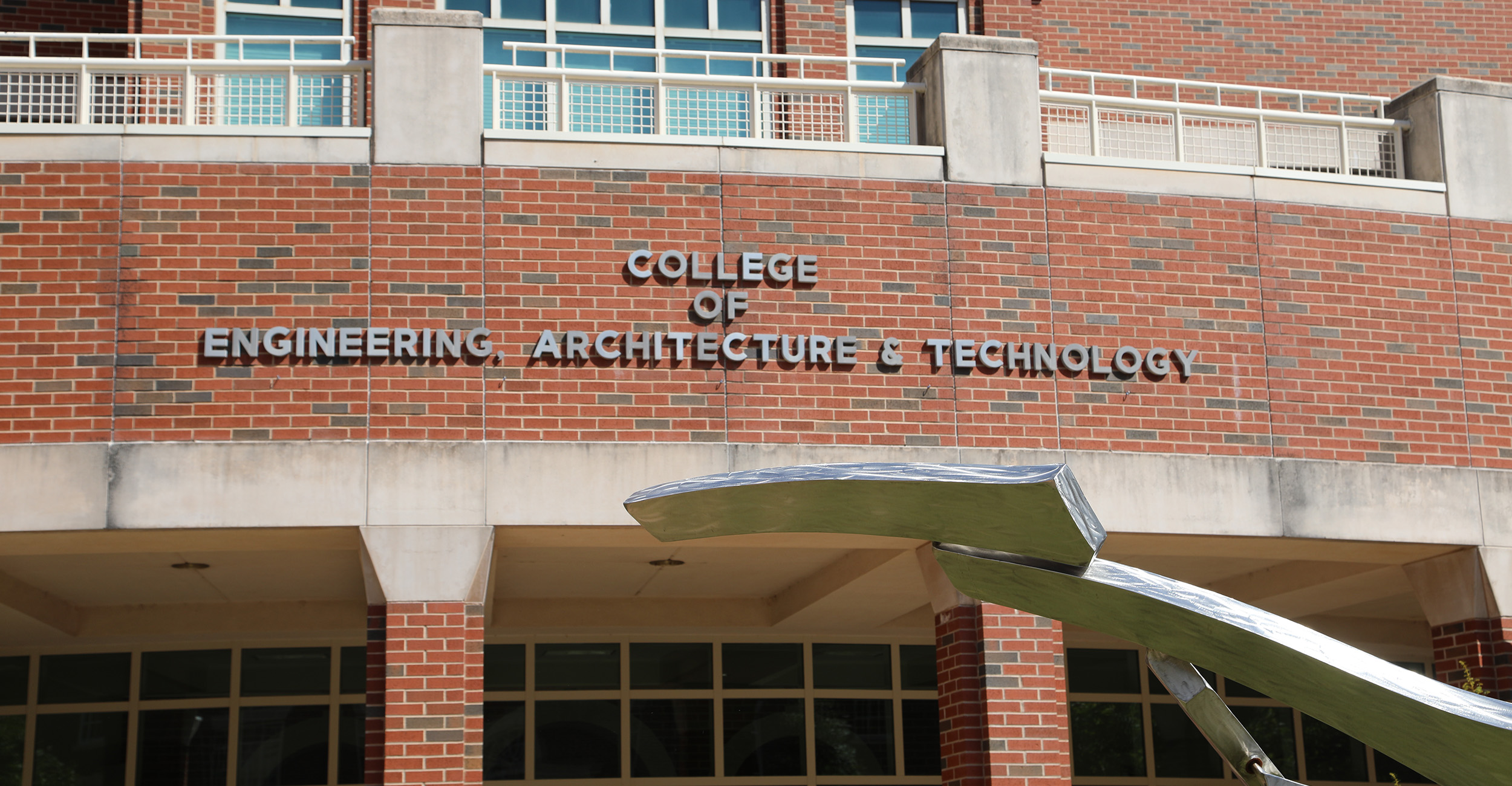 A close-up view of a red-brick academic building with large blue-tinted windows, white railings, and concrete architectural accents. Raised lettering on the brick wall reads “COLLEGE OF ENGINEERING, ARCHITECTURE & TECHNOLOGY.”