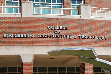 A close-up view of a red-brick academic building with large blue-tinted windows, white railings, and concrete architectural accents. Raised lettering on the brick wall reads “COLLEGE OF ENGINEERING, ARCHITECTURE & TECHNOLOGY.”