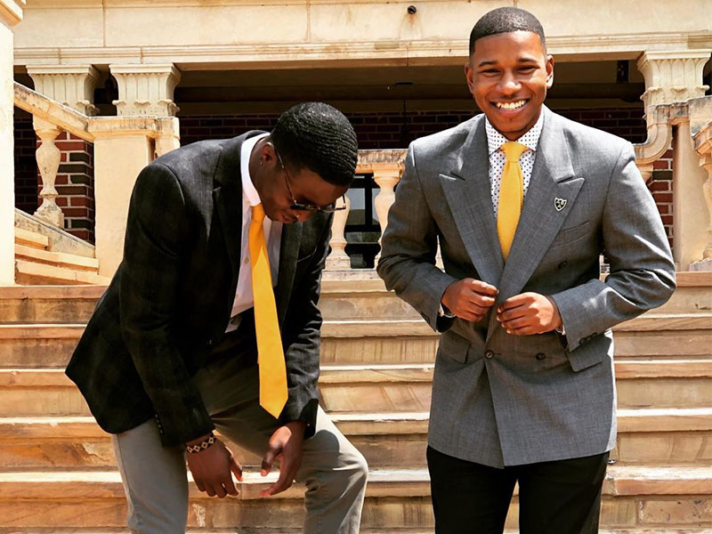 Two graduates wearing formal attire stand on stone steps outside a campus building, posing during a graduation photo session with campus architecture visible behind them.