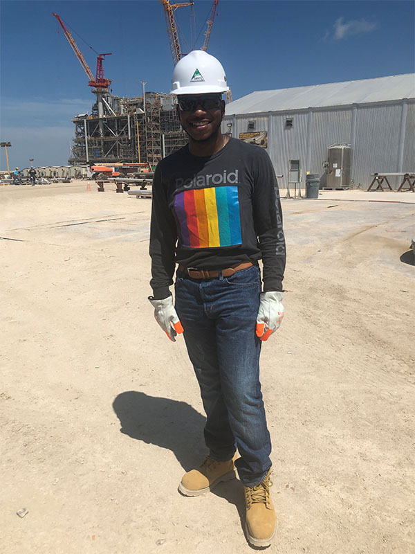 A person wearing a hard hat, gloves, and work boots stands at an industrial construction site with cranes and large structures visible in the background, representing hands‑on experience in an engineering or construction setting.