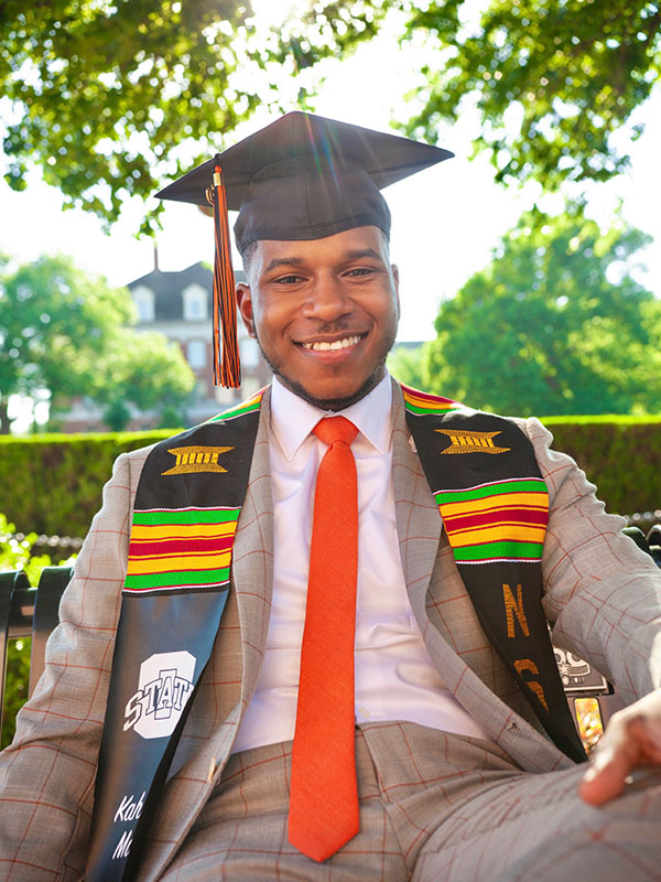 Graduate wearing a cap, stole, suit jacket, and red tie sits outdoors on campus in a formal graduation portrait, with trees and university buildings visible in the background.