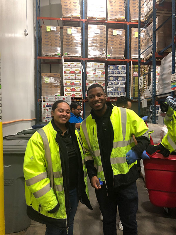 Two people wearing high‑visibility safety jackets stand inside a warehouse with stacked shelves of boxed food behind them, representing participation in a food distribution or warehouse volunteer setting.
