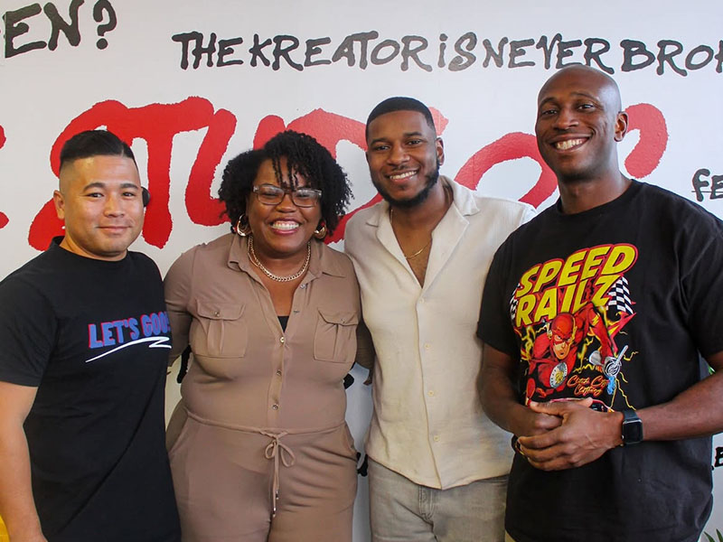 Four adults stand side by side posing for a photo in front of a wall mural with large painted text and graphics, suggesting a group or community gathering.