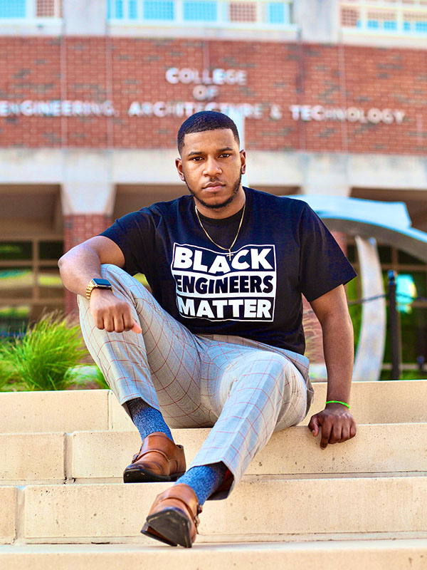Person wearing a “Black Engineers Matter” T‑shirt sits on stone steps in front of a campus engineering building, posed for a portrait highlighting engineering identity and advocacy.