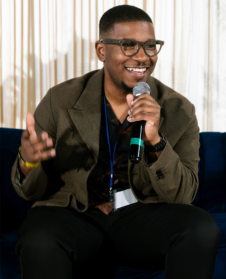 A person seated on a stage holds a microphone while speaking during a panel discussion or public talk, wearing glasses and a lanyard in an indoor event setting.