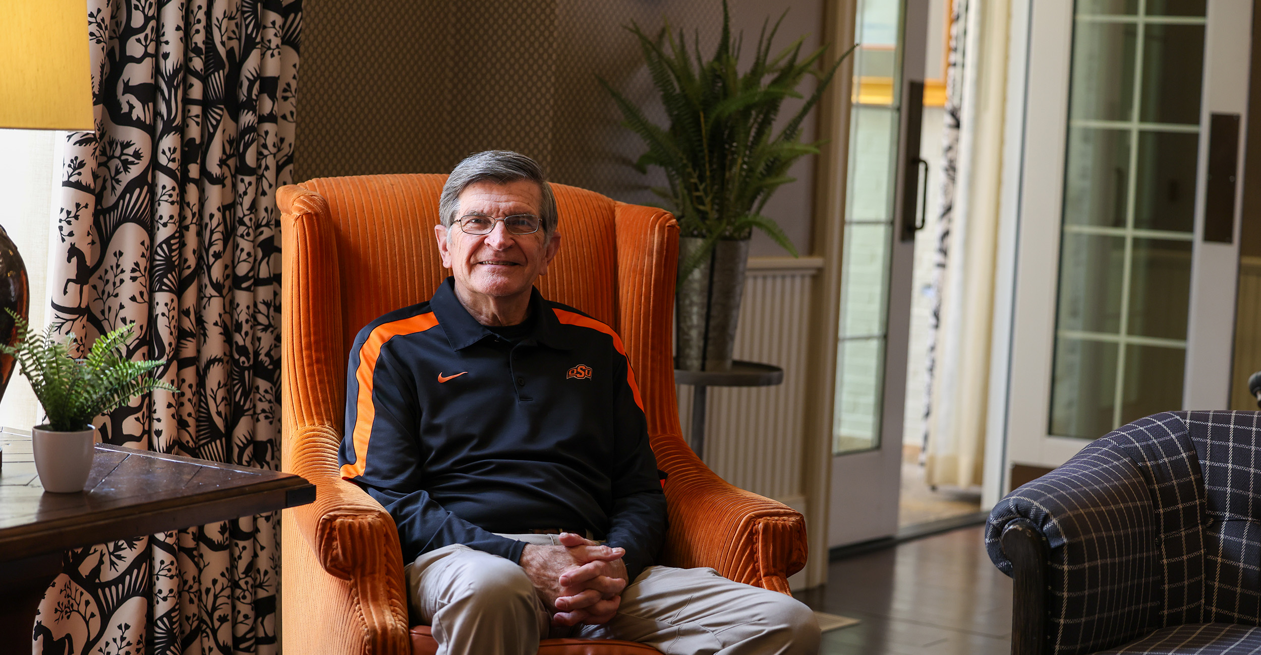 A person wearing a black-and-orange athletic pullover sits in an orange upholstered chair in a well-lit living room with patterned curtains, houseplants, and glass doors visible in the background, suggesting a posed indoor portrait.