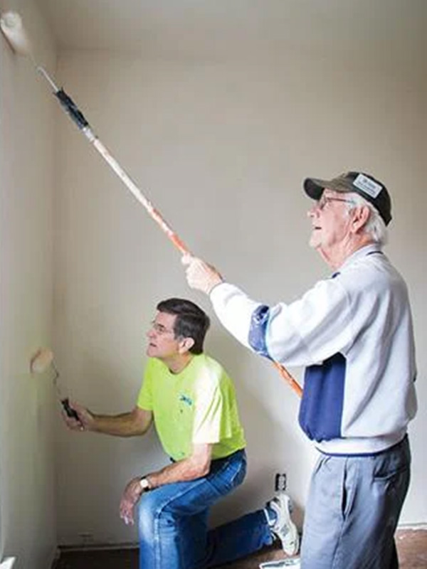Two people paint the interior walls of an unfurnished room using rollers, one standing with an extension pole and the other kneeling near the corner, illustrating hands-on volunteer or home improvement work.