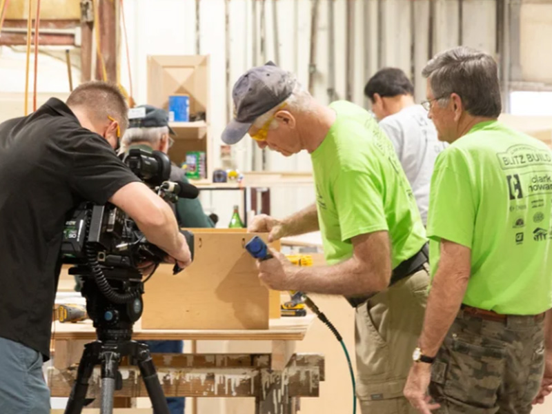 Several people in a workshop build a wooden box at a workbench while a camera operator films the process, highlighting hands-on craftsmanship and documentation of a construction activity.