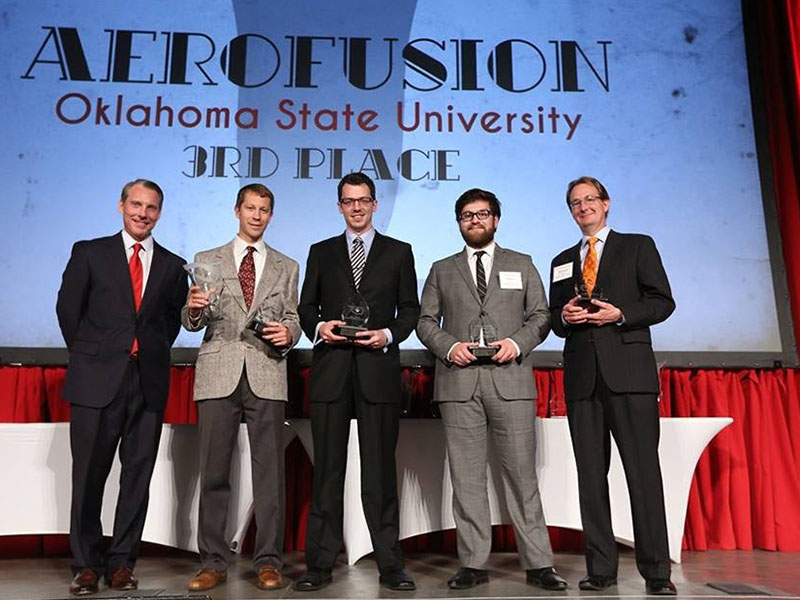 Five people in suits stand on a stage holding awards beneath a screen reading “Aerofusion Oklahoma State University 3rd Place.”
