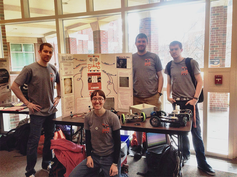 Students present a robotics project at an indoor display table with a poster board and small wheeled robots.