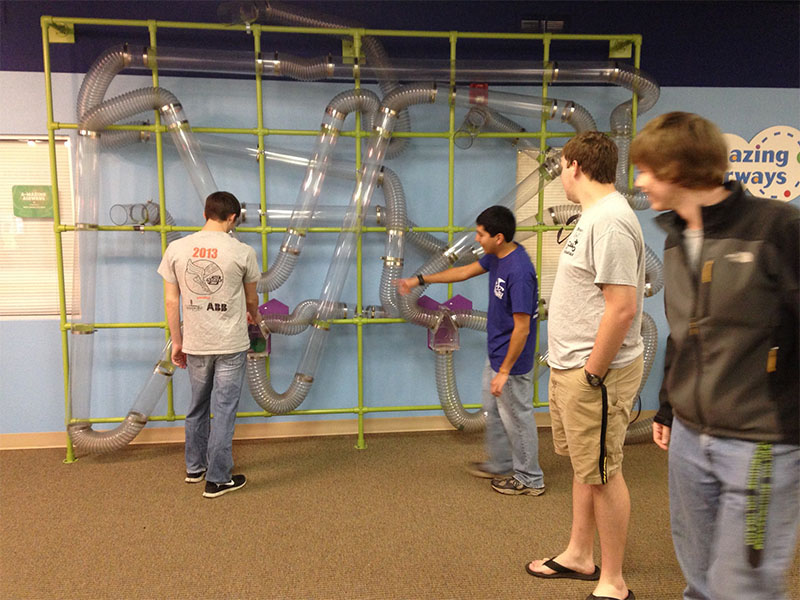 Students interact with a wall-mounted network of clear tubes and air pathways in a science museum-style exhibit
