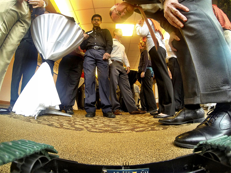 Low-angle view of people gathered around a small robot on a carpeted floor at an indoor event or demonstration
