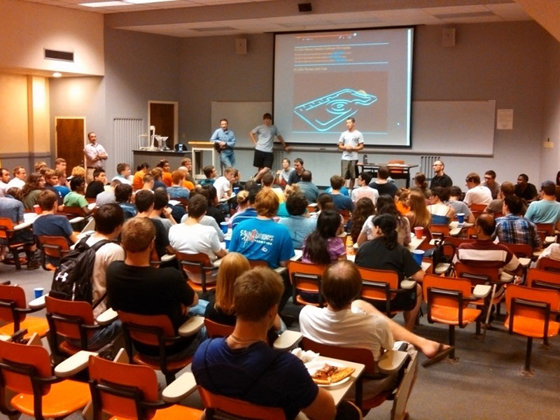 Students seated in a lecture hall watch a presentation with a projected slide showing a technical diagram.