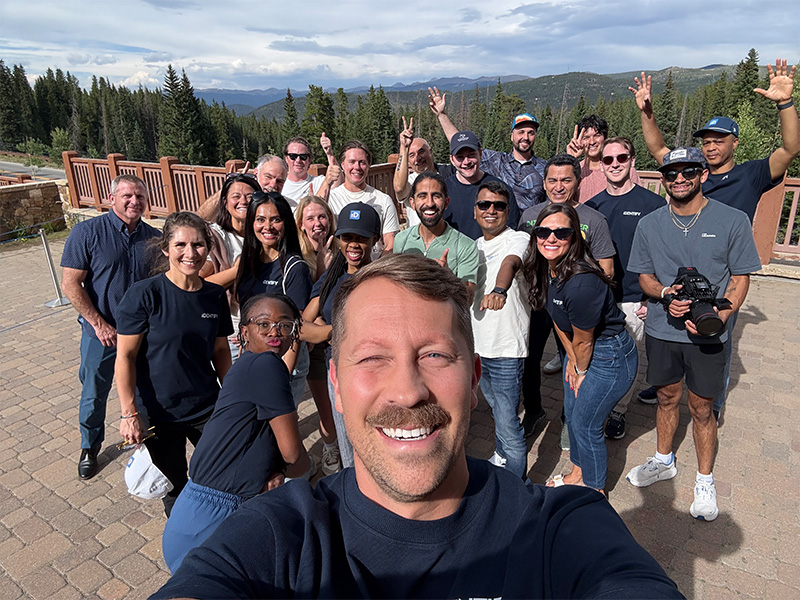 Large group of people gathered outdoors on a paved overlook, taking a group selfie with a forested mountain landscape in the background.