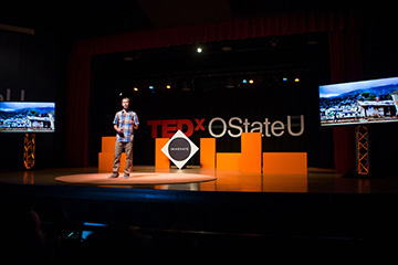 Speaker stands on a stage with a large “TEDxOStateU” sign and presentation screens on either side.
