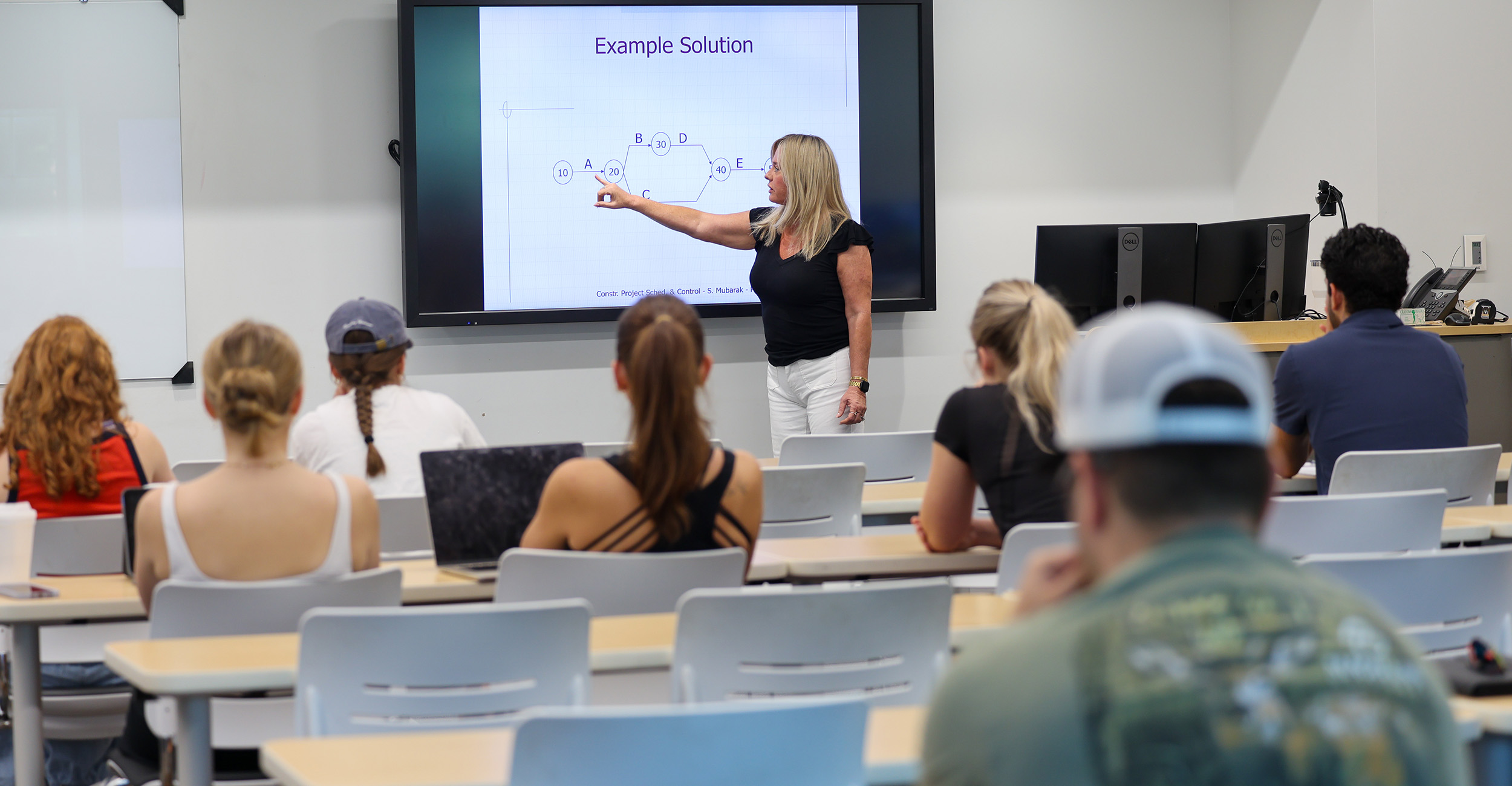 Instructor points to a diagram displayed on a large screen while students sit in a classroom lecture, following along with the lesson.