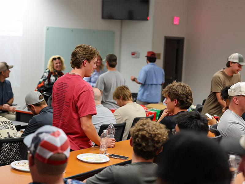 Students gather in a classroom during a group activity or break, with participants talking and moving between tables while refreshments are visible.