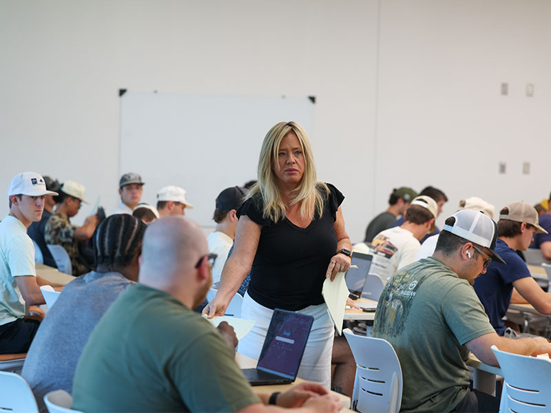 Instructor walks among students seated at classroom tables, distributing papers and providing assistance during an active learning session.