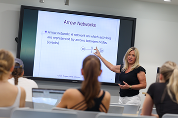 Instructor explains a network diagram on a large presentation screen while students watch from classroom seating.