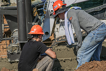 The image depicts two workers engaged in a construction project, focusing on pouring and leveling concrete.