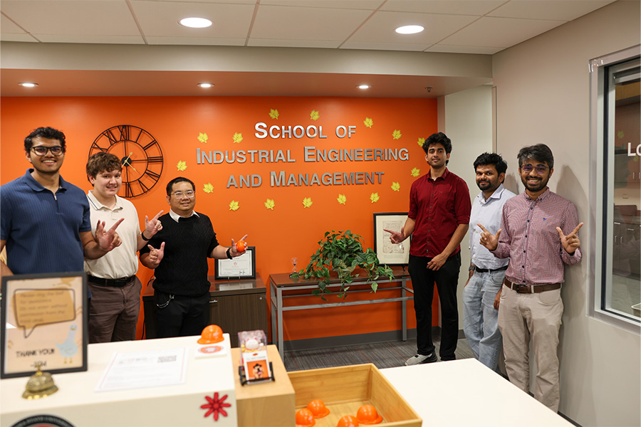 A group of six people pose inside a lobby in front of a bright orange wall displaying large metallic letters that read “SCHOOL OF INDUSTRIAL ENGINEERING AND MANAGEMENT.”