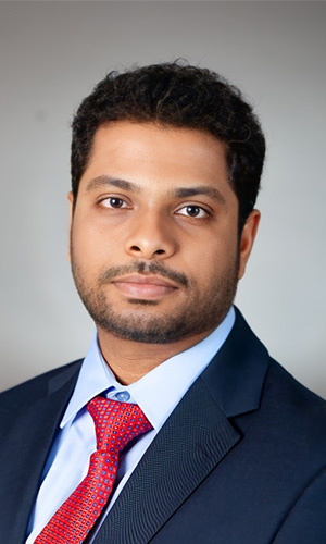 A formal head-and-shoulders portrait shows a person wearing a dark suit jacket, white dress shirt, and red patterned tie against a plain light background, suitable for a professional profile or academic bio.