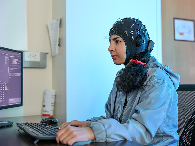 A person wearing a wired brain‑sensing cap sits at a desk typing on a keyboard while viewing code and data on a computer monitor, illustrating hands‑on research involving brain‑computer interface or robotics development.
