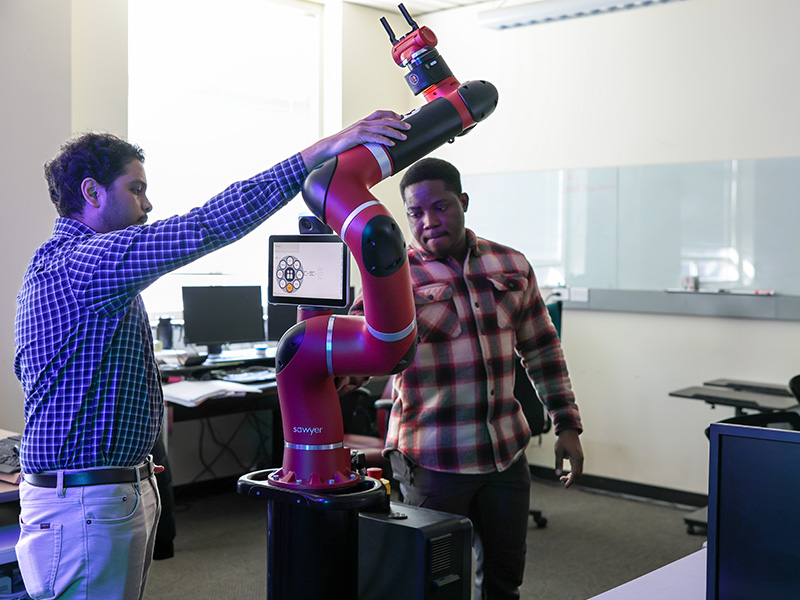 Two people adjust and guide a red collaborative robot arm in a laboratory or classroom setting, illustrating hands-on robotics research and human–robot interaction.
