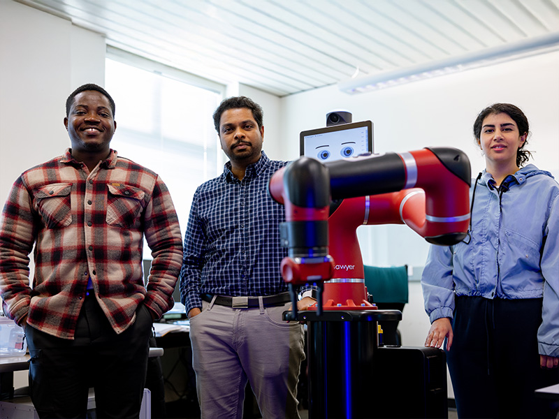 Three people stand in a laboratory or classroom beside a red collaborative robot with an attached display screen, illustrating a team-based robotics research or demonstration environment.